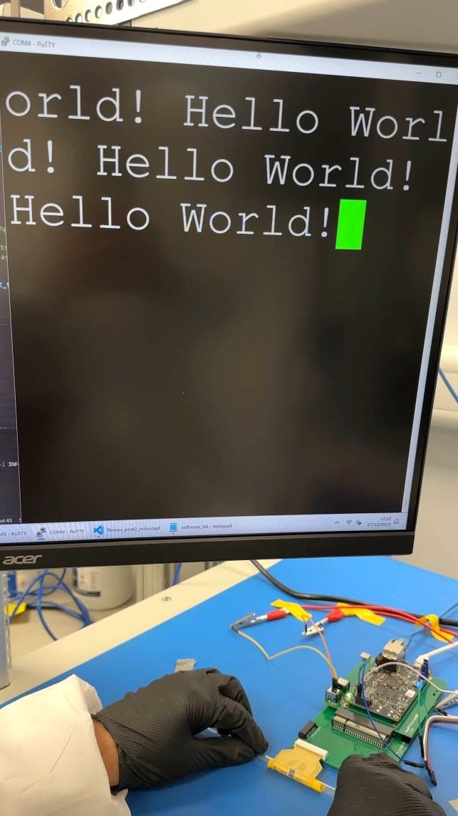 A computer monitor saying Hello World! hangs above gloved hands sitting next to a green board with a golden chip bent around a tube.