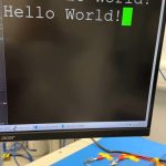 A computer monitor saying Hello World! hangs above gloved hands sitting next to a green board with a golden chip bent around a tube.