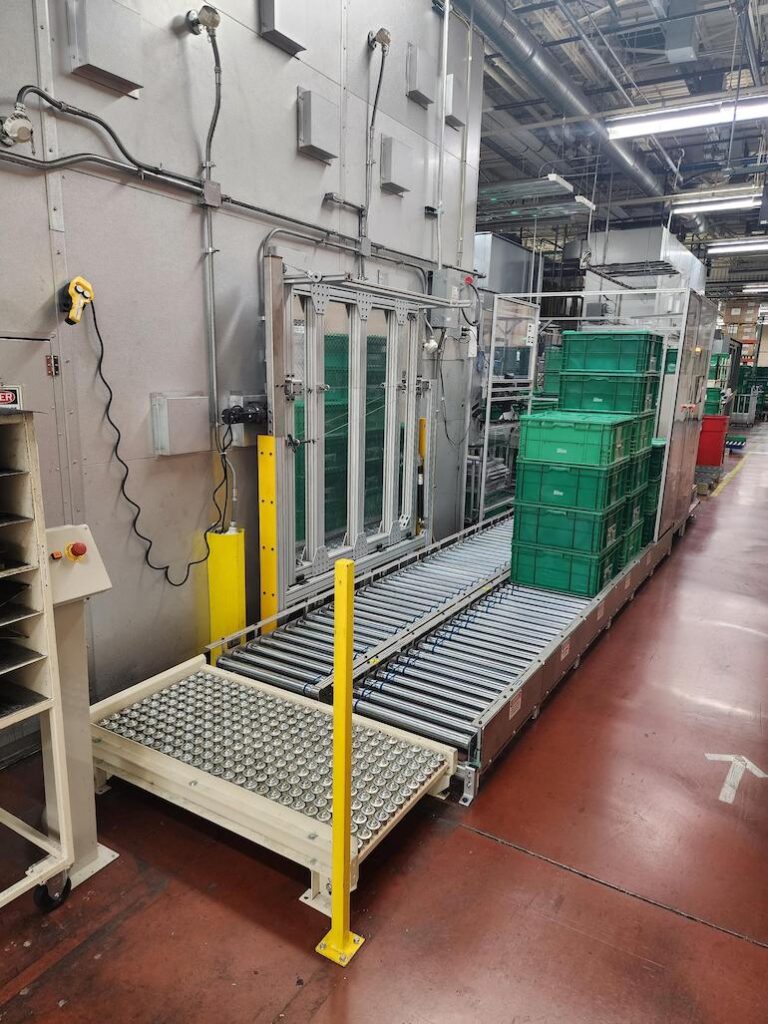 Large, heavy totes filled with automotive parts are carried to and from a paint booth on a conveyor, where a cobot picks and place totes as part of an automated process.